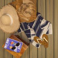 A straw sunhat, striped towel, flip-flops, and a mesh bag are arranged on a wooden deck, evoking a relaxed summer vibe. A book titled 'Funny Story' by Emily Henry sits on a wooden stool, suggesting leisure reading. The scene is bright and inviting, with neutral and navy tones dominating the palette.