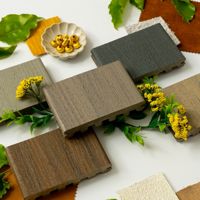 A selection of wood texture samples is arranged in a circular pattern, surrounded by colorful fabric swatches and green leaves. Yellow flowers and a small dish containing golden capsules add decorative elements to the composition. The scene is set on a white background, highlighting the natural and earthy tones of the materials.