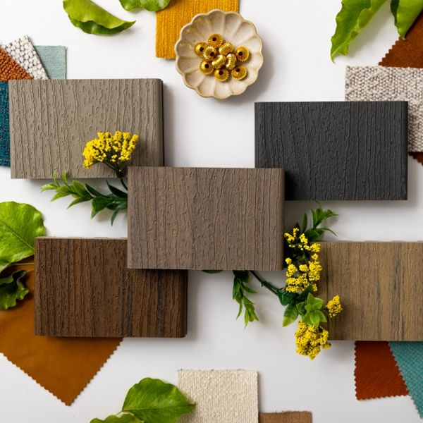 A collection of six wood texture samples is arranged on a white surface, surrounded by colorful fabric swatches in shades of teal, mustard, rust, and cream. Green leaves and yellow flowers are interspersed among the samples, adding a natural touch. A small dish with gold decorative rings sits at the top, enhancing the stylish and organic presentation.