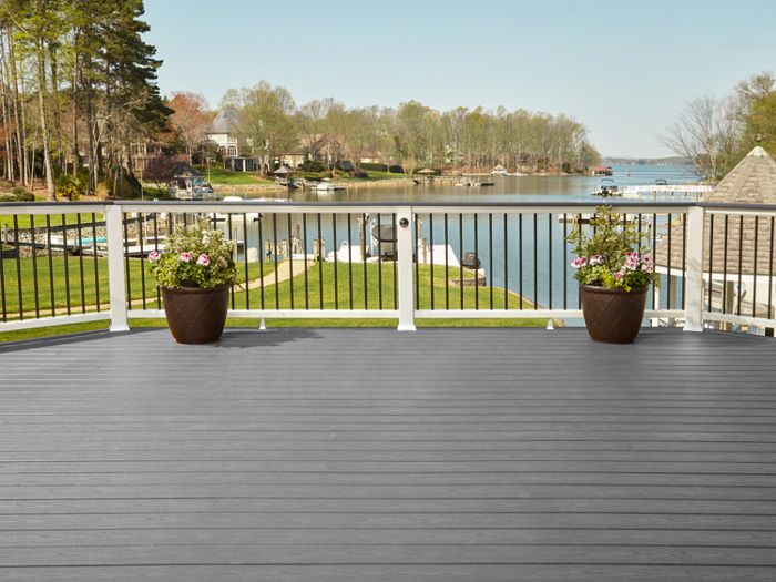 A wide, gray deck overlooks a scenic lake with houses and trees in the background. The deck features white and black railings and two large potted plants with blooming flowers. The setting is bright and tranquil, suggesting a peaceful outdoor living space with clear skies and calm water.