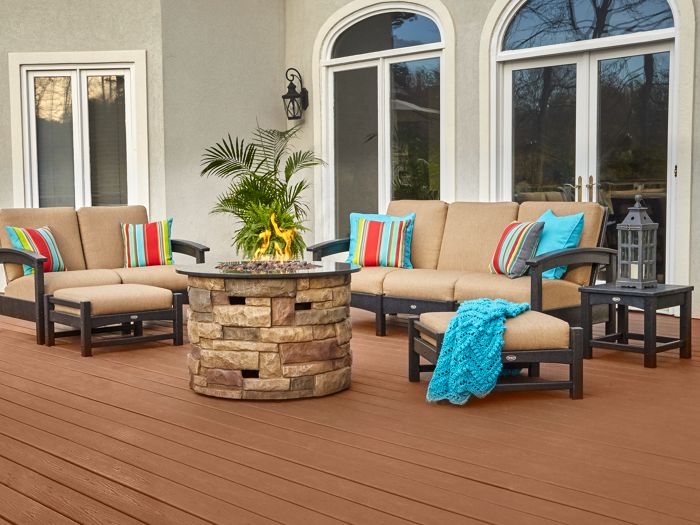 A spacious outdoor patio features cushioned seating arranged around a stone fire pit table. The setting includes a grill on the left, colorful accent pillows, and a blue throw blanket draped over a chair. Large windows and arched doors provide a view of the backyard, while potted plants add greenery to the scene. The deck is made of wide wooden planks in a warm brown tone.