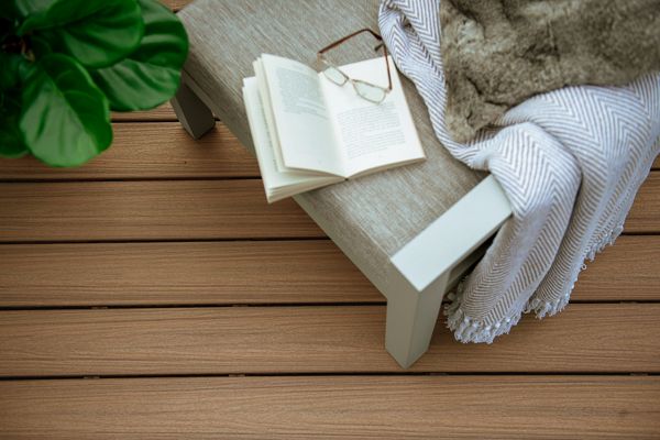 An inviting scene featuring an open book resting on a cushioned bench, accompanied by reading glasses. A soft blanket draped over the bench adds warmth to the setting, while a lush green plant enhances the natural ambiance. The wooden deck flooring completes the tranquil outdoor environment.