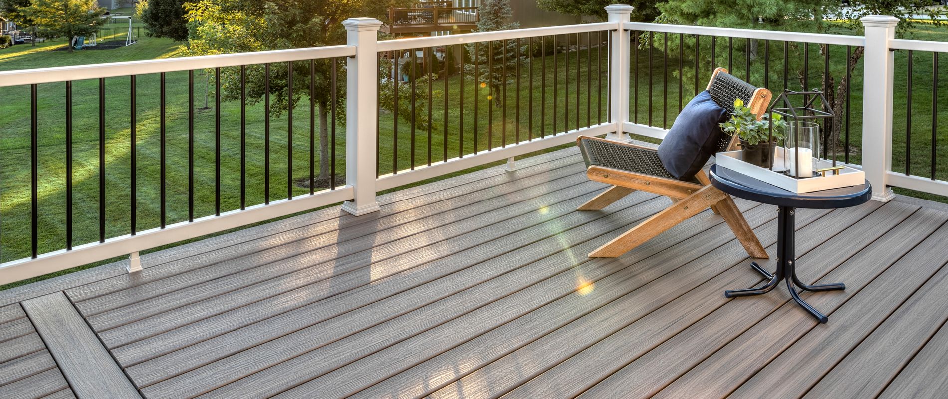 A spacious, modern deck overlooks a lush green backyard at sunset. The setting features a wooden chair with a pillow and a small round table holding a lantern and decorative items. The scene is tranquil, with warm sunlight casting long shadows and suburban homes visible in the background.