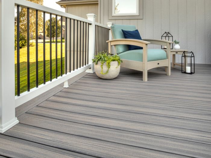 A spacious outdoor deck features wood-textured flooring and a comfortable armchair with a blue cushion. The setting includes a white and black railing, a potted plant, and decorative lanterns, creating a relaxing atmosphere. Sunlight highlights the clean lines and neutral tones of the space. The scene suggests a peaceful residential backyard or patio area.