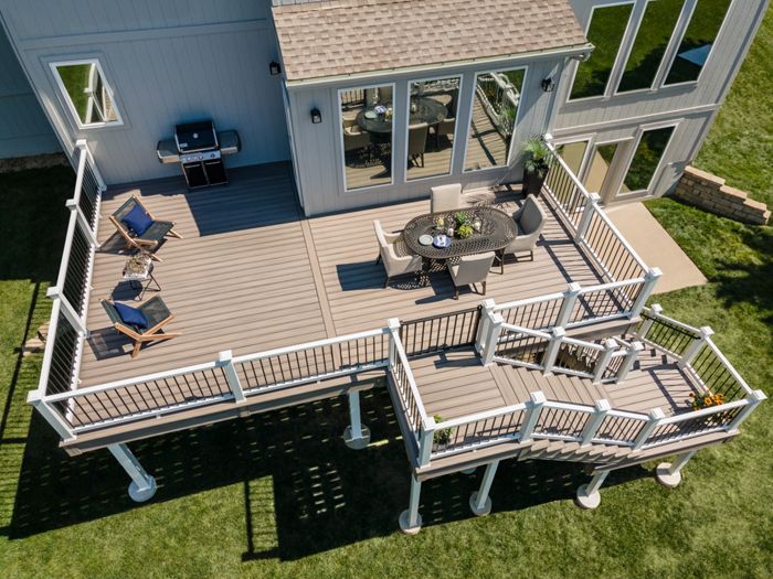 A large, elevated wooden deck is attached to a modern house, featuring a round dining table with chairs and two lounge chairs. The deck includes a staircase leading down to a grassy backyard and a grill positioned near the house wall. The setting is bright and sunny, with clean lines and neutral tones creating a welcoming outdoor living space.