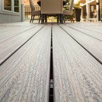 A detailed close-up view of composite deck boards showcases the wood grain texture and clean lines. The setting is an outdoor patio area with a table and chairs visible in the background, suggesting a relaxed and inviting atmosphere. Warm natural light highlights the neutral tones of the decking, creating a modern and comfortable outdoor living space. No visible text or numbers are present in the image.