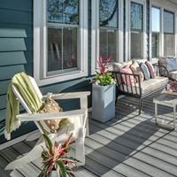 A spacious outdoor deck features comfortable cushioned seating, a white chair, and a coffee table with a vibrant flower arrangement. Potted plants add a touch of greenery, while sunlight streams across the gray deck boards. The setting is inviting and relaxed, perfect for lounging or entertaining in a residential backyard. Large windows reflect the bright day, enhancing the airy atmosphere.