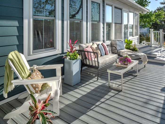 A spacious outdoor deck features a cozy seating area with a cushioned sofa, armchair, and coffee table. Decorative pillows and potted plants add color and vibrancy to the space, while sunlight streams across the gray deck boards. The setting is inviting and tranquil, ideal for relaxation or social gatherings.