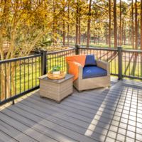 A spacious outdoor deck features a single wicker armchair with a blue cushion and orange throw, positioned beside a matching wicker side table holding a basket of fruit. The deck overlooks a sunlit forest with tall trees and golden foliage, creating a tranquil and inviting atmosphere. The scene is brightly lit, emphasizing the clean lines of the deck and the natural surroundings.