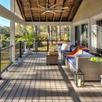 A large, covered outdoor patio features a wicker sofa with blue cushions and a cozy orange throw. The setting overlooks a lush, wooded backyard, with sunlight streaming through the trees. Decorative elements include a potted plant, a lantern, and a small coffee table, creating a relaxed and inviting atmosphere.