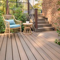 A cozy outdoor deck features a wicker chair with a blue cushion and a matching side table, set against a backdrop of lush greenery. The deck leads up to a staircase with dark railings and is adjacent to a brick house wall. Sunlight filters through the trees, creating a warm and inviting atmosphere with natural wood tones and soft shadows.