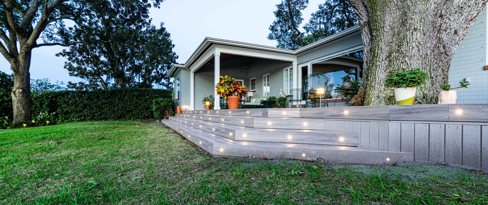 A spacious backyard features a contemporary patio area with a covered seating space and large wooden deck steps. The steps are illuminated with small built-in lights, creating a welcoming ambiance as evening falls. Mature trees frame the scene, and potted plants add a touch of greenery to the modern outdoor setting.
