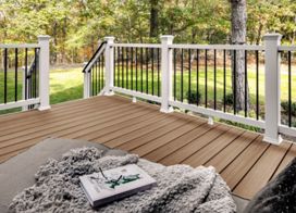 A wooden deck featuring a white and black railing surrounded by lush greenery. The setting includes a cozy blanket and a book placed on a soft cushion, suggesting relaxation. The scene is bright and inviting, with natural light filtering through the trees.