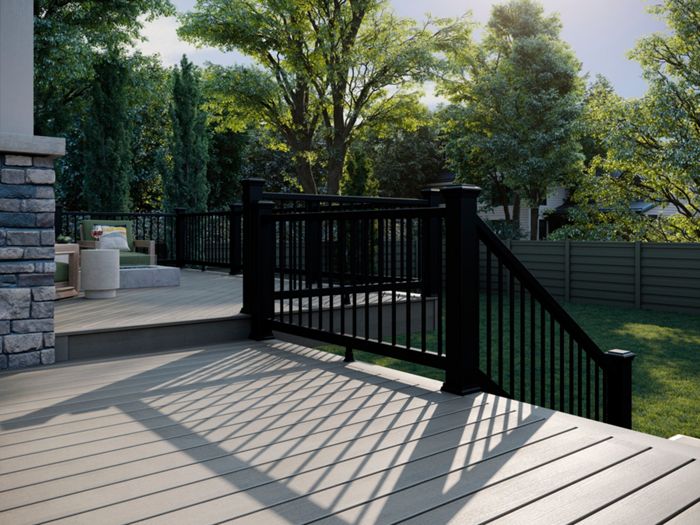 A spacious outdoor deck features sleek black railings and light wood planks, bathed in afternoon sunlight. The setting is a suburban backyard with lush green trees and a privacy fence in the background. Shadows from the railing create geometric patterns on the deck surface, adding visual interest. Outdoor seating and a stone accent wall are visible, enhancing the inviting atmosphere.