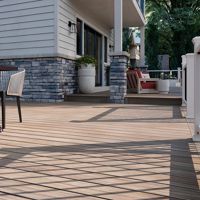 A spacious wooden deck extends from a contemporary house, featuring stone siding and horizontal paneling. The setting includes a metal dining table with chairs and a large planter, while lounge seating is visible in the background. Sunlight casts geometric shadows across the deck, creating a warm and inviting outdoor atmosphere.