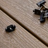 A close-up view shows several Trex fasteners and screws resting on a light brown, wood-textured composite decking. The fasteners are black with the 'Trex' name clearly visible on one of them. The setting highlights the installation process for outdoor decking, focusing on the hardware and material details. No people or additional text are present.