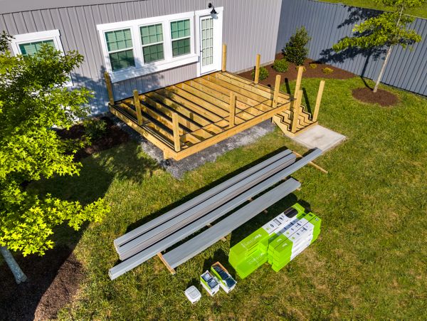 A partially built wooden deck is attached to the back of a house, with framing and posts in place. Construction materials, including planks, boxes, and tools, are neatly arranged on the grass nearby. The setting is a sunny backyard with green grass, trees, and a gray exterior wall with windows. No people are present, and the scene emphasizes outdoor home improvement. Several green and white boxes display visible text and numbers.