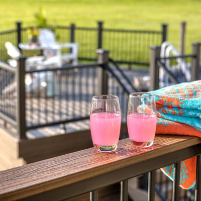 Two glasses filled with a pink beverage rest on a wooden deck railing next to a colorful towel. The background features a fenced outdoor pool area with white chairs and lush green grass. The scene is brightly lit, suggesting a sunny, relaxed atmosphere perfect for summer leisure.
