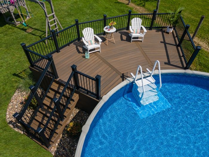 A circular above-ground pool is connected to a spacious wooden deck featuring two white Adirondack chairs and a small round table. The deck is bordered by black railings and accessed by a set of stairs, with the pool ladder leading directly into the clear blue water. The setting is outdoors on a sunny day, surrounded by green grass and landscaping stones.