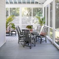 A spacious screened-in porch features a gray wooden dining table surrounded by matching chairs. The setting is airy and filled with natural light, with potted plants adding a touch of greenery. The ceiling is vaulted with exposed beams and a white ceiling fan, creating a relaxed and inviting atmosphere. The overall palette is neutral with soft gray and white tones.