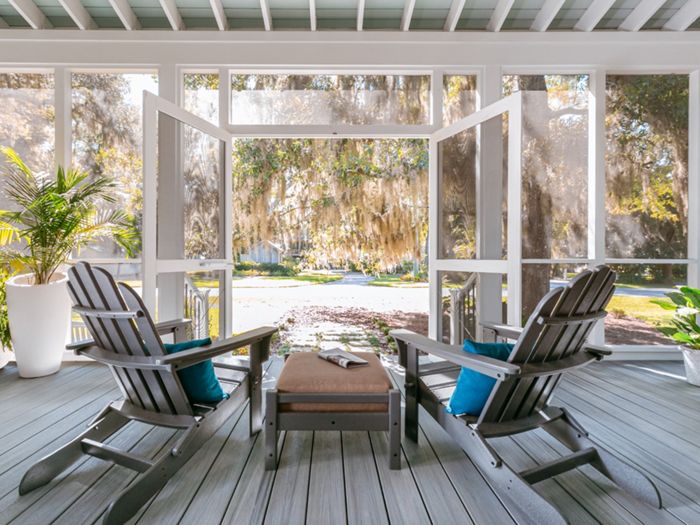 A bright, inviting screened porch features two dark Adirondack chairs with blue accent pillows facing a small table. The porch is decorated with lush green potted plants and overlooks a sunlit outdoor landscape with hanging moss. The open double doors enhance the airy, relaxed atmosphere, and the wooden floorboards add a touch of rustic charm.