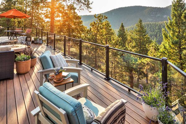 A spacious wooden deck overlooks a lush, sunlit forest and distant mountains. Comfortable lounge chairs with blue cushions and a small table create a relaxing seating area, while potted plants add a touch of greenery. The glass railing provides an unobstructed view of the natural landscape, and an orange umbrella shades a dining table in the background. Warm sunlight enhances the tranquil and inviting atmosphere.