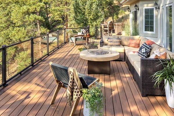 A spacious wooden deck features contemporary wicker patio furniture, including a sectional sofa and chairs arranged around a round fire pit table. The setting is surrounded by glass railings and overlooks a lush, forested landscape. Decorative pillows, a throw blanket, and potted plants add a cozy, inviting touch to the scene. The area is bathed in natural sunlight, creating a warm and relaxed atmosphere.
