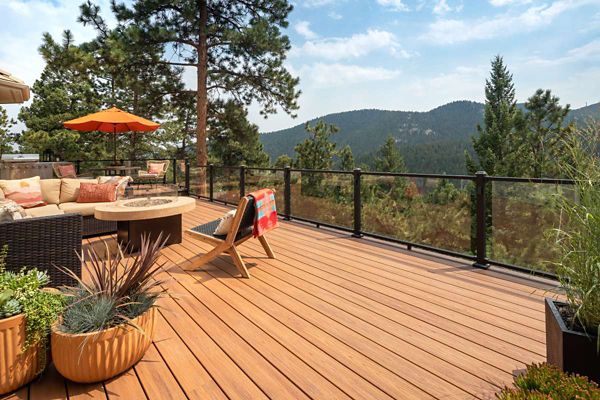 A large wooden deck overlooks a scenic mountain landscape, surrounded by tall pine trees. The setting features comfortable outdoor furniture, a fire pit table, and an orange patio umbrella, creating a cozy and inviting atmosphere. Potted plants add greenery, while glass railing panels provide unobstructed views of the natural surroundings. The overall mood is relaxed and tranquil, ideal for outdoor gatherings.