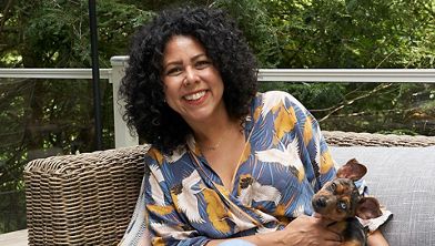 A woman with curly hair sits on a wicker outdoor sofa, holding a small brown and black dog on her lap. She is wearing a colorful patterned blouse and blue jeans. The setting appears to be a patio or garden area with greenery in the background, creating a relaxed and casual atmosphere.