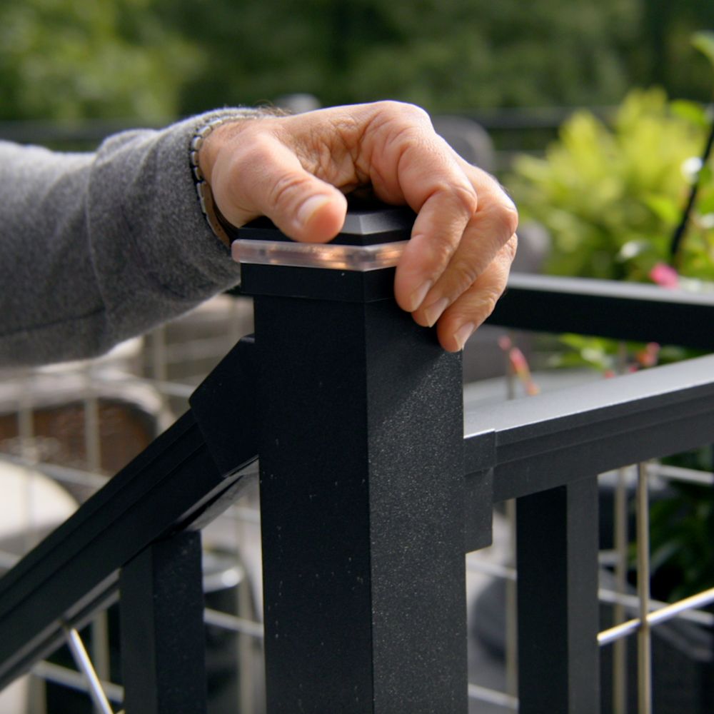 A close-up of an adult hand resting on a black Trex deck railing post, with a garden or patio setting in the background. The scene features greenery, potted plants, and a wire fence, suggesting an outdoor leisure space. The Trex logo is visible on the lower right, confirming the product brand. The lighting is natural, and the mood is calm and inviting.