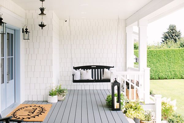 A welcoming farmhouse-style porch features a black wooden swing with white cushions, set against white shingle siding. The space is decorated with potted plants, a large black lantern, and a tan welcome mat with a sunburst design. The porch overlooks a manicured lawn and is accented by classic black lantern light fixtures. The overall mood is airy, inviting, and peaceful.