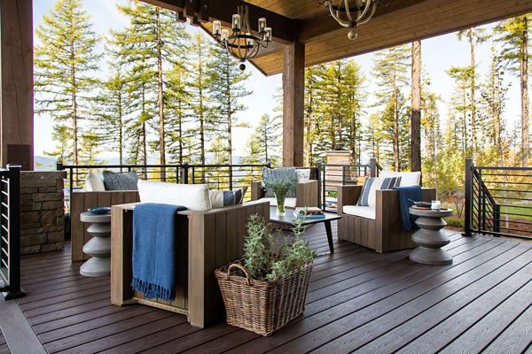 A contemporary outdoor patio features wooden armchairs with white cushions and blue accent throws. The setting overlooks a lush forest, creating a tranquil and inviting atmosphere. Decorative elements include a woven basket with greenery, round side tables, and elegant lighting fixtures. The space is designed for relaxation and social gatherings, with natural light enhancing the cozy ambiance.