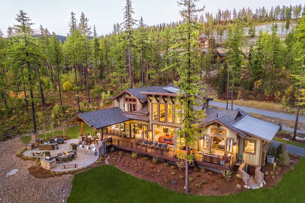 A contemporary multi-level house is nestled among tall pine trees in a forested mountain setting. The home features expansive windows, a spacious wooden deck, and a well-lit outdoor seating area. Warm interior lighting creates a cozy atmosphere, while the surrounding landscape includes manicured lawns and natural stone pathways. The scene is captured at dusk, highlighting the tranquil and inviting mood.