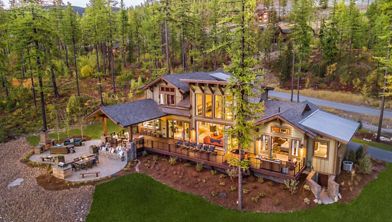 A contemporary multi-level house is nestled among tall pine trees in a forested mountain setting. The home features expansive windows, a spacious wooden deck, and a well-lit outdoor seating area. Warm interior lighting creates a cozy atmosphere, while the surrounding landscape includes manicured lawns and natural stone pathways. The scene is captured at dusk, highlighting the tranquil and inviting mood.