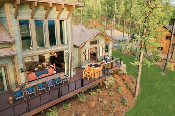 A contemporary house with large windows and an expansive deck is nestled in a forested mountain setting. The deck features multiple seating areas and a round hot tub, overlooking a landscaped hillside. The interior is visible through open doors, showcasing a cozy living space with natural light and wood accents.