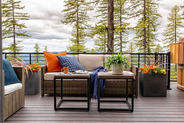 A comfortable outdoor patio features a cushioned sofa with colorful pillows and a throw blanket, set against a scenic backdrop of tall pine trees and mountains. The setting includes modern black tables, a ceramic mug, and a potted plant centerpiece, with vibrant flowers in large planters adding a lively touch. The overall mood is inviting and tranquil, ideal for relaxation or enjoying a warm beverage outdoors.