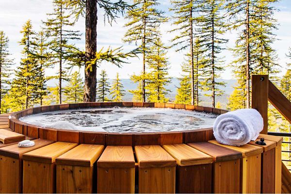 A round wooden hot tub filled with bubbling water is set on a deck surrounded by tall pine trees. A neatly rolled white towel rests on the edge of the tub, suggesting relaxation and comfort. The scene is bathed in natural daylight, with a tranquil forest backdrop and warm wood tones. No visible text or numbers are present in the image.