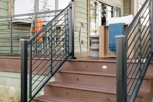 A set of brown wooden stairs with sleek black metal railings leads up to a contemporary deck. The deck features a wooden outdoor chair with a white cushion and a blue throw blanket, set against a green-paneled house. Reflections in the windows and a bright orange accent add visual interest to the modern outdoor setting.