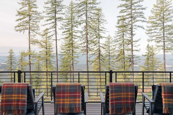 Four deck chairs draped with colorful plaid blankets are arranged on a wooden terrace. The setting overlooks a scenic pine forest with tall, slender trees and distant mountains visible through the railing. The atmosphere is tranquil and cozy, with a soft, natural color palette and a focus on outdoor relaxation.