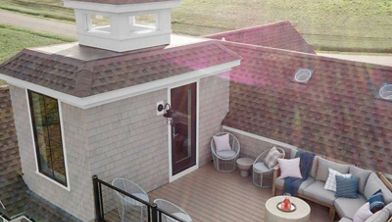 A stylish rooftop patio features a sectional outdoor sofa, round coffee table, and accent chairs arranged for relaxation. The setting is atop a residential building with shingle roofing, surrounded by a cable railing for safety. The area is bathed in natural light, with green fields visible in the background, creating a serene and inviting atmosphere. Neutral tones and blue accents contribute to a calm, contemporary mood.