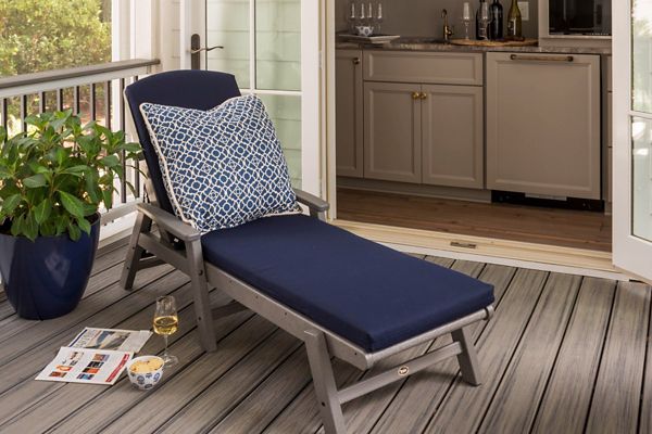 A navy blue cushioned lounge chair with a patterned pillow is positioned on a wooden porch deck. The setting includes a potted plant, a glass of white wine, a cup, and a magazine placed nearby, suggesting a relaxing atmosphere. The porch opens into a kitchen area with cabinets and glassware visible inside. The overall mood is calm and inviting, ideal for leisure or reading.