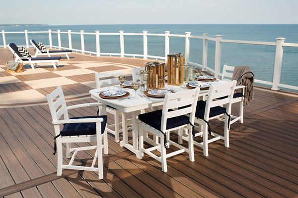 A white wooden dining table with matching chairs is arranged on a spacious deck overlooking the ocean. The table is set with plates, glasses, and metallic pitchers, suggesting a meal or gathering. In the background, lounge chairs with striped cushions face the water, creating a relaxed and inviting atmosphere. The setting is bright and airy, with a coastal, nautical style and a neutral color palette.