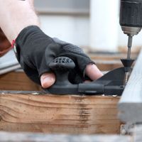 A close-up view shows a gloved hand operating a power drill to fasten a screw into a wooden plank. The setting appears to be an outdoor construction or renovation project, likely involving deck installation or repair. The image highlights the use of protective gloves and a drill guide for precision. The color palette features natural wood tones and muted grays, emphasizing a practical, work-focused atmosphere.