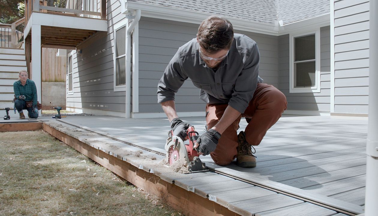 A man is kneeling on a wooden deck, using a circular saw to trim the edge of the boards. The setting is an outdoor residential backyard with a modern gray house in the background. Another person is seated further back, observing the activity. The scene conveys a sense of home improvement and DIY craftsmanship, with natural daylight and neutral tones.