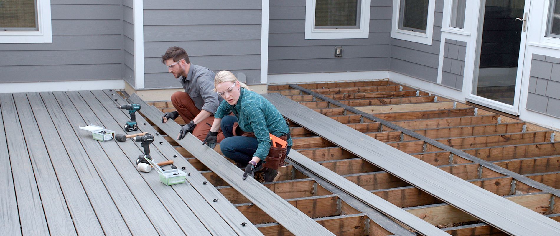 A man and woman are working together to install composite deck boards on a partially completed patio. The setting is a residential backyard with gray siding and multiple windows visible. Tools and materials are spread around as they align and secure the decking. The mood is industrious and collaborative, with neutral and gray tones dominating the scene. No visible text or numbers are present in the image.