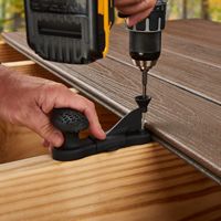 A close-up view shows a person using a power drill to fasten a composite deck board onto a wooden frame. The scene takes place outdoors, with autumn foliage visible in the background. The image highlights the use of a specialized deck fastener and the detailed texture of the decking material. The person's hands are visible, focusing on the precise installation process.