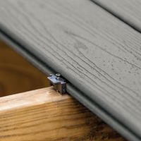 Close-up view of a composite deck board being installed onto a wooden frame using a hidden fastener system. The image highlights the textured surface of the gray deck board and the metal fastener securing it in place. The natural wood grain of the support structure contrasts with the synthetic decking material. No visible text or numbers are present in the image.