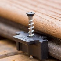 A black deck fastener with a silver screw is shown securing two wooden planks. The image focuses on the installation process, highlighting the fastener's placement between the boards. The setting appears to be an outdoor deck, with natural wood textures and warm lighting. No visible text or numbers are present on the fastener or screw.