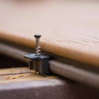 A detailed close-up image shows a metal screw fastened through a black plastic clip, securing a wooden board in place. The setting appears to be a woodworking or construction environment, with the focus on the hardware and wood grain. The image highlights the installation process and the precision of the materials involved. No visible text or numeric values are present in the image.