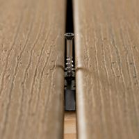 A detailed view shows a screw securing two composite deck boards together. The image focuses on the narrow gap between the boards, highlighting the wood grain texture and precise installation. The setting suggests a construction or home improvement project, with neutral brown tones dominating the scene. No visible text or numbers are present.